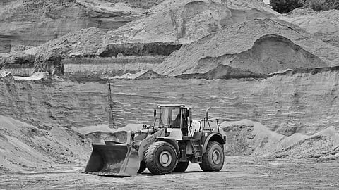 A yellow front loader in a quarry, surrounded by layered sand and gravel, under a clear sky. The scene conveys industriousness and ruggedness.