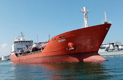 Large red cargo ship named "Darmik" sailing in calm waters under a clear blue sky. Other vessels and a shoreline are visible in the background.
