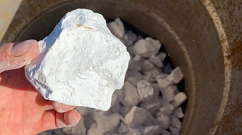 A hand holds a large white rock over a rusty metal container filled with similar rocks, under bright sunlight, creating a rugged, earthy tone.