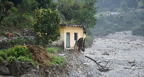 A broken house on the edge of a mostly-dry riverbed, with trees on the left.