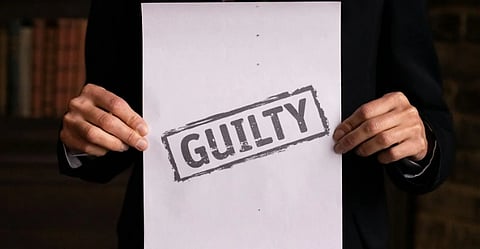 A person in a suit holds a paper with "GUILTY" stamped on it in bold letters. The background features a brick wall and bookshelves, suggesting a legal setting.