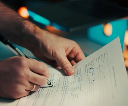 Image of hands holding and signing a document with a pen, set on a desk with a blurred laptop in the background, conveying a sense of focus and professionalism.
