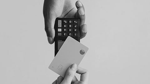 A hand holds a small payment terminal as another hand reaches up with a credit card. The background is gray, conveying a neutral, modern tone.