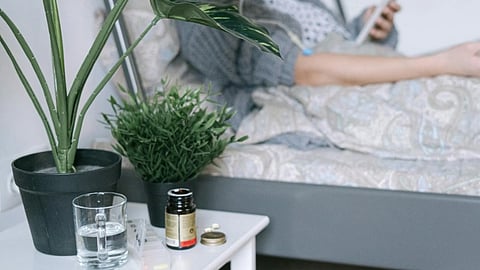 Person resting in bed, holding a phone; nearby, a table with a plant, a glass of water, and medication bottles, conveying a calm, recovering atmosphere.