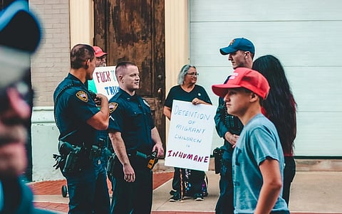 A protest scene with two police officers and several demonstrators holding signs about migrant detention. The mood is tense and serious.