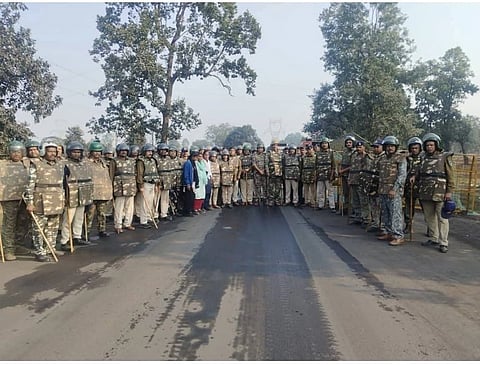 A group of police personnel standing in the middle of a road in riot gear, with a few trees in the background.
