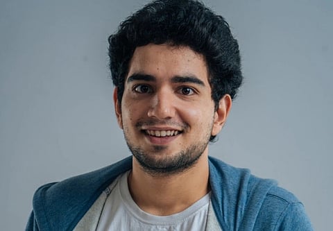 Image of stand-up comedian Samay Raina wearing a white shirt with a blue graphic and a blue hoodie. Background is plain gray.