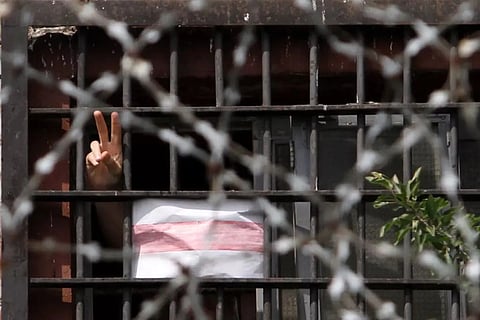 A photo of a jail cell through barbwire, with a hand reaching through, holding up the peace sign, and a free belarus sign next to it.
