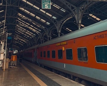 Image of a train platform under an arched glass and metal roof. An orange train is stationed on the right, with platform number 5 signage visible. The scene is calm and orderly.