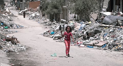 A young girl in a red outfit in Gaza walks down a debris-filled street, carrying a small appliance. The scene conveys desolation and resilience amid urban rubble.