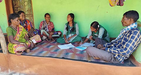 Children sitting on a mat, talking to elders and taking notes in front of a green wall.