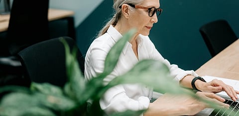 A focused woman with gray hair and glasses types on a laptop in an office. She wears a white blouse. A green plant is in the foreground.