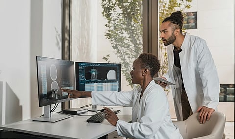 Two scientists in lab coats analyze medical scans on dual monitors. One points at the screen, while the other observes with interest, holding a tablet.