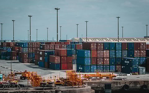 A busy port scene with stacked colorful shipping containers, primarily red and blue. Orange machinery and tall light poles are visible under a clear blue sky.