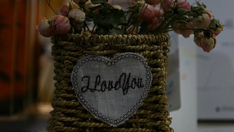 A woven basket holding dried pink roses sits on stacked books. A heart-shaped patch reads "I love you," conveying a warm, affectionate tone.