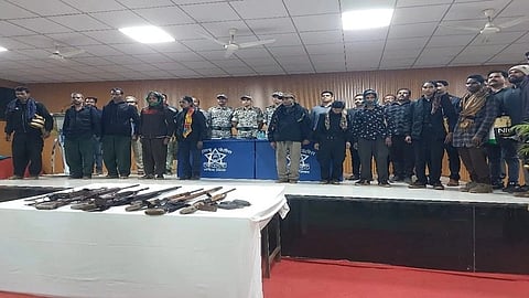 A group of Maoists stand on a stage in front of CRPF officers, with weapons laid out on a table in front of them.