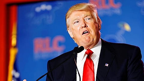 Trump in the middle of giving a speech in 2014. There are two mics in front of him and a blue board behind him with the acronym 'RLC'.