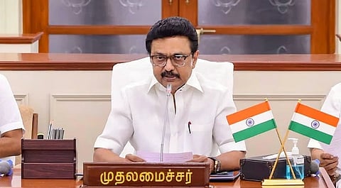 Image of Tamil Nadu Chief Minister M. K. Stalin in a white shirt sits at a desk with two small Indian flags. He appears focused, speaking into a microphone, suggesting a formal meeting setting.