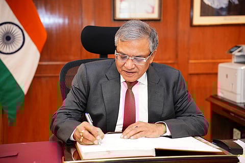 Chief Election Commissioner Gyanesh Kumar signing ledger in his office. He is wearing a suit. There is a wooden wall behind him and an Indian flag to the left.