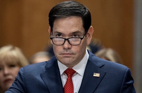 Image of Secretary of State Marco Rubio in a blue suit and red tie stands, looking focused. He wears glasses, and a U.S. flag pin is on his lapel. Blurred audience in the background.