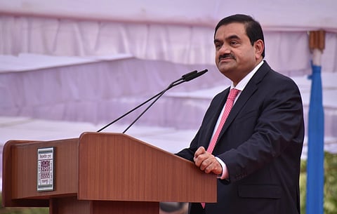 Gautam Adani in a dark suit and red tie speaks at a podium with a microphone. The setting is formal, with a calm and professional atmosphere.
