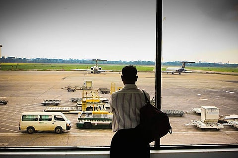 A man with a back looks out at a runway through a window. There is a van nearby and two planes in the distance.