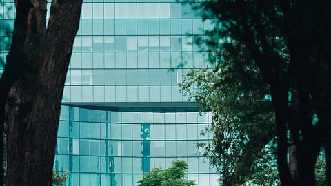 Image of Tall trees frame a sleek, modern glass building with a reflective facade, creating a serene cityscape blending nature with urban architecture.