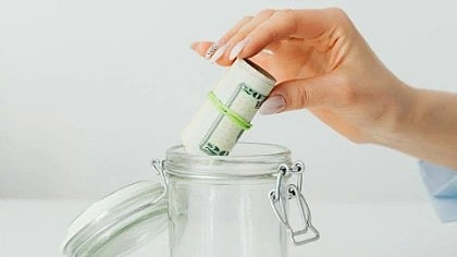 A hand places a rolled bundle of cash into an open glass jar on a white surface, conveying themes of saving money and financial planning.