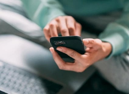 A person in a green sweater sits cross-legged on a bed, using a smartphone. A partially visible laptop is nearby, suggesting multitasking and focus.