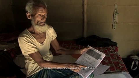 An old survivor of the Nellie massacre sits in front of the camera on his bed, pointing at a document. A subtitle reads "These are my documents from 1935-"
