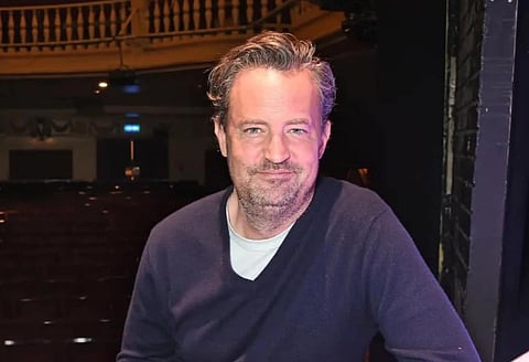 Image of late Friend's actor Mathew Perry. He is wearing a blue sweater. the background of the image includes seats on stairs depicting a studio like theatre room