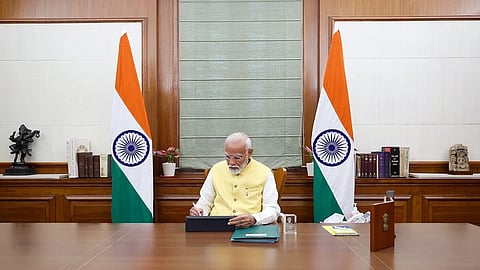 Prime Minister Narendra Modi sitting in his office and working. There are two flag each on one side of the chair he is seated on.