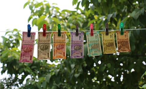 Image of Indian banknotes of various denominations clipped onto a clothesline, set against a background of lush green trees, suggesting outdoor freshness.