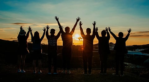 Seven silhouetted people stand on a hill, arms raised in joy against a vibrant sunset, conveying a sense of unity and celebration.