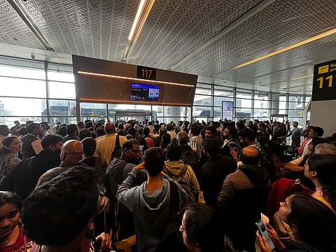 Hundreds of passengers wait at the Hyderabad airport due to Indigo flight cancellations. The passengers are waiting in the lobby area, with glass windows in their front.