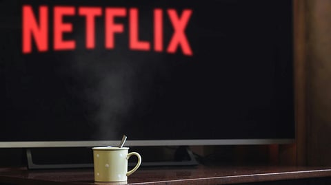 A cozy scene with a steaming mug on a table in the foreground, and a TV in the background displaying the red Netflix logo.