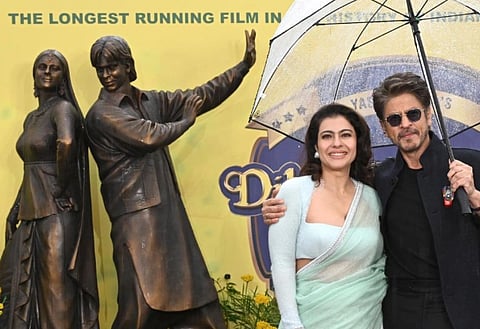 Image of Shah Rukh Khan and Kajol in London to unveil their Bronze statue. They are dressed elegantly, stand smiling under an umbrella beside bronze statues of themselves. The backdrop mentions a long-running film.