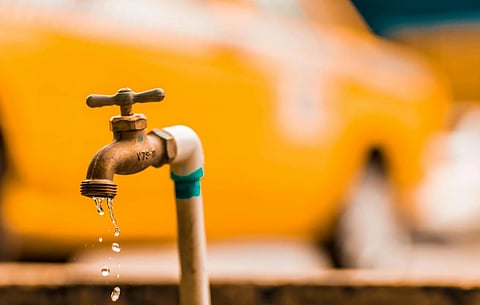 Close-up of a dripping outdoor faucet with blurred yellow car in the background. The droplets emphasize a sense of urgency and waste.