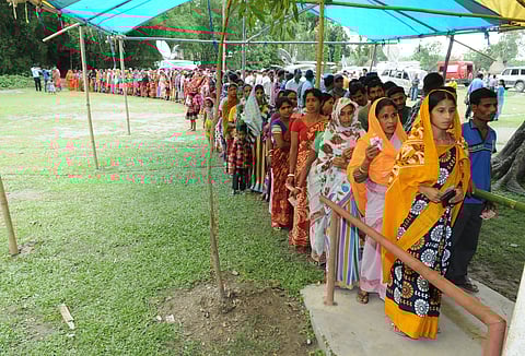 A queue of female voters are seen standing at a polling booth. Next to the female queue, a queue of male voters is there. The location is an open ground, green grass on the land and trees behind, with a tent where the voters are standing.