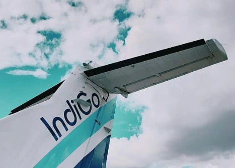 The image shows the tail of an IndiGo aircraft against a vibrant sky with clouds. The logo is visible, conveying a sense of travel and freedom.