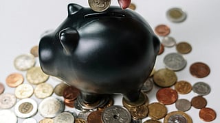 A hand places a coin into a black piggy bank surrounded by scattered coins on a white surface. The image conveys a theme of saving and financial growth.