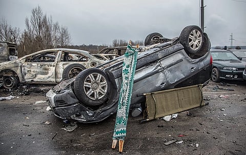 The image depicts Overturned car on a bleak, debris-covered road with a prayer rug and cot leaning against it. Burned vehicles in the background convey a scene of destruction. Image is of Bucha after Russian invasion of Ukraine