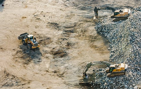 Aerial view of a construction site with three excavators operating on rocky, uneven terrain. Two excavators are on the right, moving debris, conveying industrious activity.
