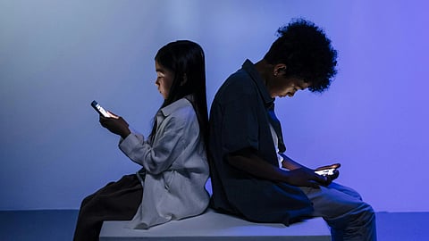Two people sit back-to-back on a low block, absorbed in their smartphones. The room is dimly lit with a blue hue, creating a contemplative mood.