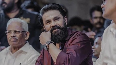 In the image Malayali Actor dileep is shown. he is wearing a shirt and his hand is on his maustache the background looks like an event