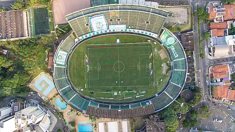 Aerial view of a large, empty soccer stadium surrounded by city buildings and trees. The field is green with clear markings.