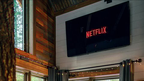 Cozy cabin interior with large windows, a wooden desk, red stools, and a screen displaying Netflix. A dog sits on a red cushioned bench.