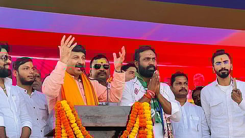 Manoj Tiwari speaks energetically at a podium adorned with orange marigold garlands, surrounded by a group of men in white. The tone is lively and vibrant.