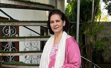 Image of Navjot Kaur Sidhu in a pink outfit with a white scarf stands smiling in front of a spiral staircase with greenery.