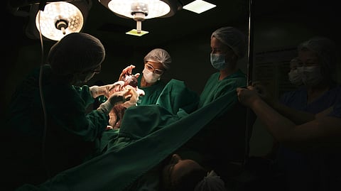 Doctors in surgical attire hold a newborn under bright lights in a dimly lit operating room during a C-section, conveying a moment of new life and care.